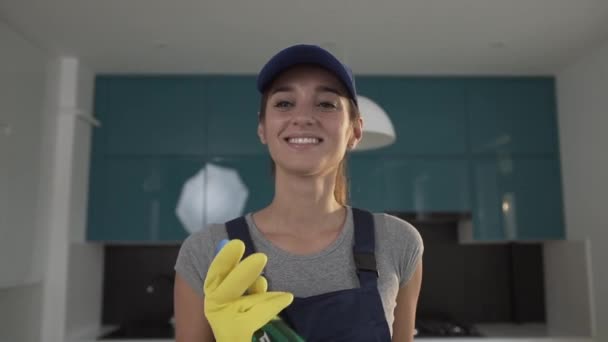 Schöne junge Putzfrau in Arbeitskleidung, die mitten in der modernen Küche grünes Waschmittel versprüht — Stockvideo