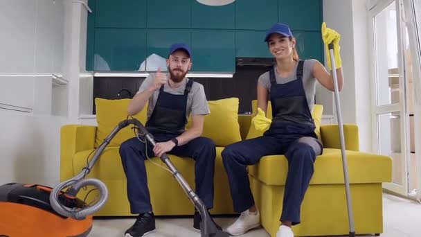 Mosolygó fiatal férfi és női csapata a takarítási szolgáltatást ül a konyhában kanapé és a mutató remek gesztusok — Stock videók