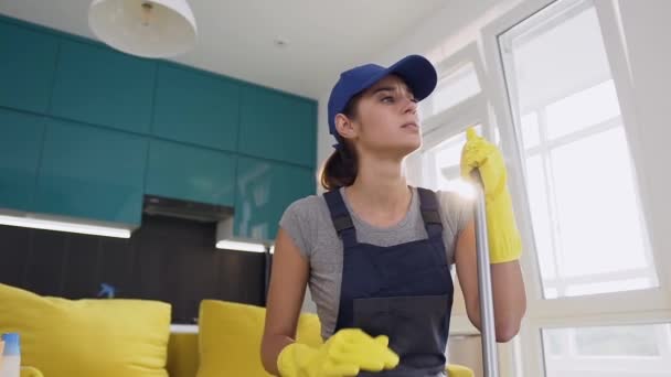 Jovencita triste del servicio de limpieza con guantes de goma amarillos sosteniendo una fregona y sentada en el cómodo sofá en medio de la cocina — Vídeo de stock