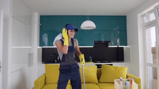 Laughing pretty woman in earphones from cleaning service listening the music and dancing with a mop in the kitchen — Stock Video