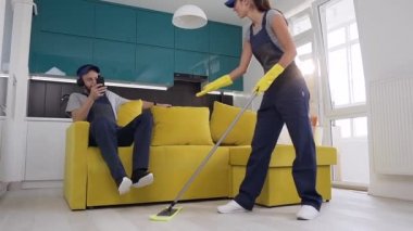 Temizlik servisi kızgın güzel kadın ahşap zemin wipping ve eşi kanepede oturan ve kulaklıkla müzik dinlerken