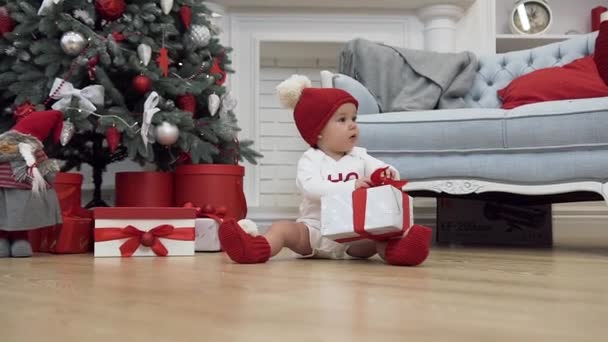 Aantrekkelijk beeld van glimlachende baby in kerst kleding zittend op de houten vloer en spelen met een geschenk — Stockvideo