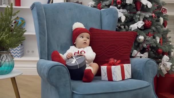 Imagen atractiva de la niña dulce en ropa de Navidad sentado con regalos de año nuevo en silla cómoda cerca del árbol de Navidad — Vídeo de stock