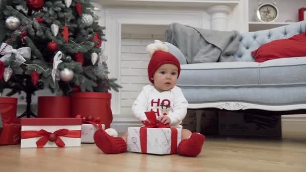 Dulce bebé en sombrero de santa sentado cerca del árbol de Navidad de hadas y tratando de abrir el regalo de Navidad — Vídeos de Stock