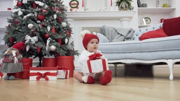 Foto di Natale di un bambino dolce in abiti natalizi seduto sul parquet e con in mano un regalo vicino all'albero di Natale — Video Stock