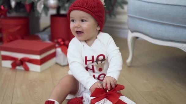 Bonito bebê sorridente em roupas engraçadas ano novo siiting perto da árvore de Natal e jogando com um belo presente — Vídeo de Stock