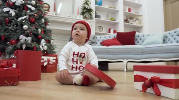 süß lächelndes Kleinkind, das Spaß mit dem Deckel des Kastens hat, der auf dem Holzboden neben dem wunderschönen Weihnachtsbaum sitzt