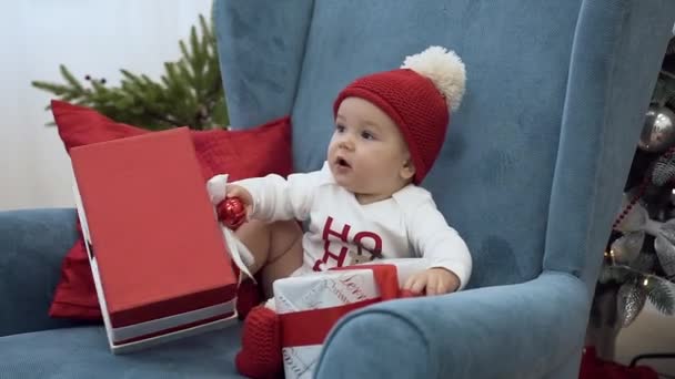 Báječný batole sedí ve velkém křesle a hraje si s vánočními dárky. — Stock video