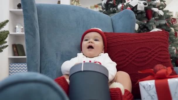 Adorable bebé sonriente con gran regalo sentado en un gran sillón de brazo azul con almohada roja cerca del árbol de Navidad — Vídeo de stock