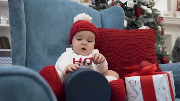 Sweet little baby girl having fun with christmas present sitting in comfortable arm chair near christmas tree — Stock Video
