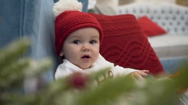 赤い帽子の愛らしい幼児のクローズアップ白いボブルは、枕と大きな椅子に座って、贈り物を楽しんでいます — ストック動画