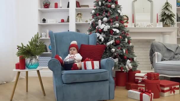 Красивый милый малыш сидит в большом синем кресле с подушкой среди рождественских подарков в празднично украшенной рождественской комнате — стоковое видео