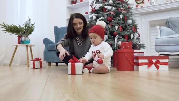 Mãe bonita sentada no chão perto da árvore de natal com seu bebê adorável que tenta pegar bola vermelha — Vídeo de Stock
