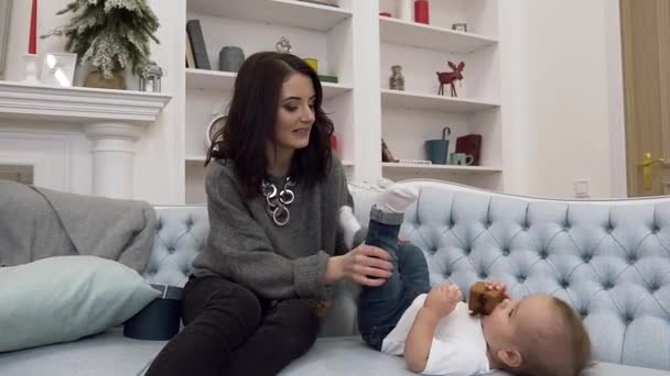 Улыбающаяся красавица-мама играет с детскими ножками, пока лежит на удобном диване — стоковое видео