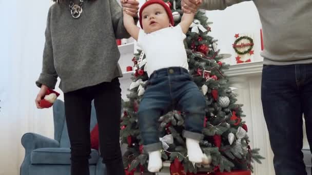 Felice uomo e donna sorridente tenendo da entrambe le mani il loro bambino carino e alzare le mani in alto sullo sfondo dell'albero di Natale — Video Stock