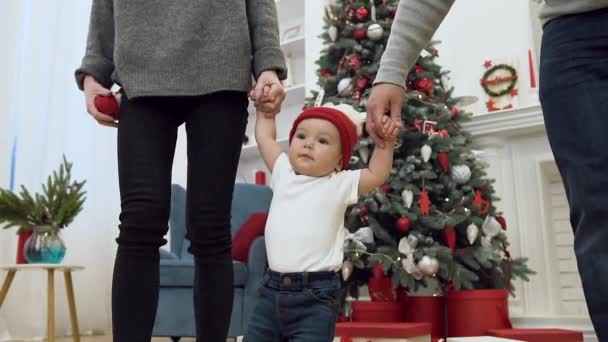 Mouvement lent de l'incroyable mignon tout-petit qui tient par les deux mains de ses parents et marche près de l'arbre de Noël — Video