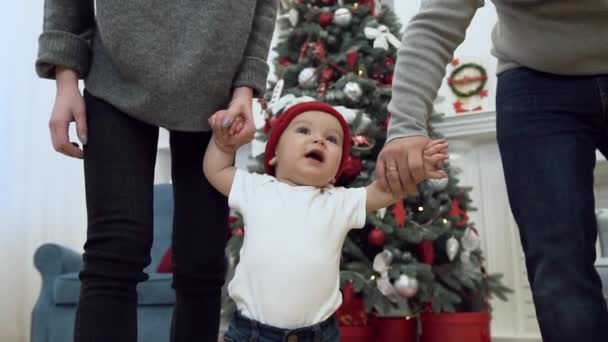 Απίστευτο χαριτωμένο νήπιο προσπαθεί να περπατήσει ενώ οι γονείς του κρατώντας τον από τα δύο χέρια στο εορταστικό δωμάτιο με χριστουγεννιάτικο δέντρο — Αρχείο Βίντεο