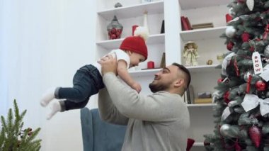 İyi görünümlü sevgi dolu baba noel ağacı ile odada onun inanılmaz sevimli gülümseyen bebek havada kusuyor