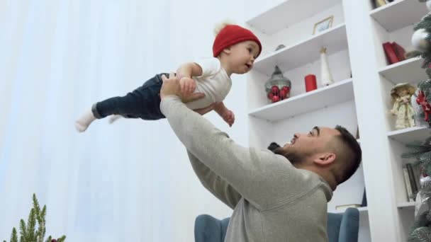 Красивый любящий отец держит своего милого младенца в воздухе в комнате с красиво украшенной елкой — стоковое видео