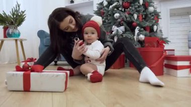 Yeni yıl hediyesi kırmızı topu düzeltmek istiyor onun sevimli annesi ile noel ağacı yakınında yerde oturan bobble şapka sevimli bebek