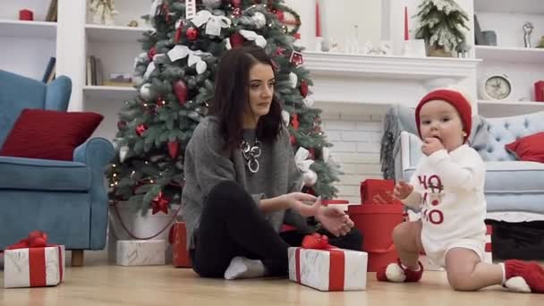 Beautiful young woman sitting on the floor near christmas tree with her cute baby girl which gnawing red ball — Stock Video