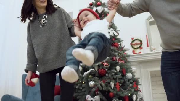 Encantadora família sorridente segurando seu bebê bonito por ambas as mãos e levantá-los no alto no fundo da árvore de Natal — Vídeo de Stock