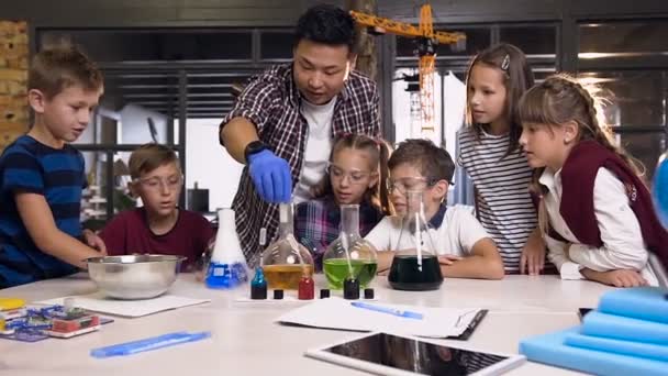 Učitel asijských věd s dětmi v laboratorní třídě pracuje na experimentu tím, že se suchý led vloží do baněk s barevnou tekutinou, zatímco v obou lahvemi byla reakce kouře ze suchého ledu — Stock video