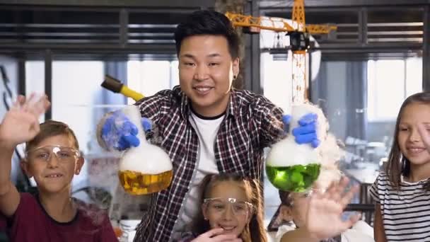 Retrato de un grupo de seis alegres jóvenes estudiantes caucásicos saludando con las manos mientras su maestro coreano sostiene dos flaks con líquido de colores donde el humo frío se evapora — Vídeo de stock