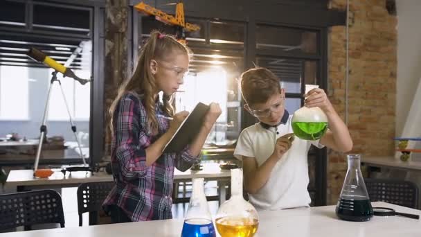 Two young scholars of caucasian pupils in safety goggles doing experiment with green liquids in flask and dry ice — Stock Video
