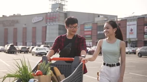 Χαρούμενο ελκυστικό ασιατικό ζευγάρι που ωθείται μπροστά τους ένα κάρο γεμάτο από διάφορα προϊόντα — Αρχείο Βίντεο