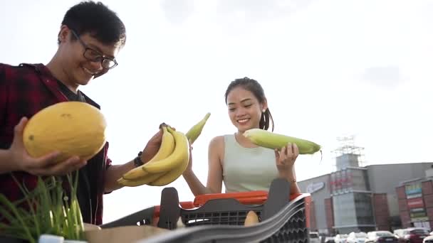 Feliz joven asiática chica sosteniendo en sus manos maíz y guapo chico guardando melón y babanas — Vídeos de Stock