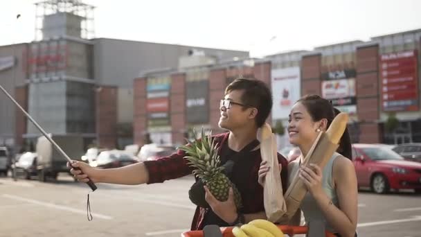 Funny young vietnamese pair of man and woman making selfie with peanapple and long bread near the supermarket — Stock Video