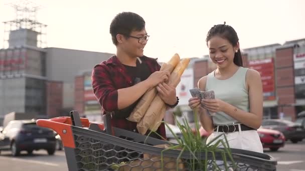 Красивий 30-й в'єтнамський хлопець у окулярах Холдинг сендвіч ролика і його мила подруга підрахунку грошей біля великого магазину — стокове відео