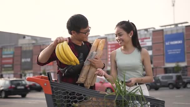 Attraktives Bild eines lustigen asiatischen Paares, das mit einem Marktwagen voller Lebensmittel steht und miteinander scherzt — Stockvideo