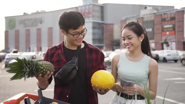 Ймовірно усміхнений азіатський чоловік важить в руках арахіс і диню і його гарненька дівчина намагається вгадати, що важче — стокове відео