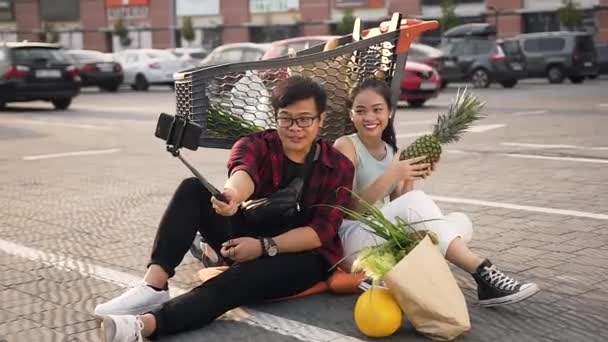 Satisfecho joven asiático pareja sentado en el suelo cerca de mercado trolley y hacer selfie con manzana — Vídeo de stock