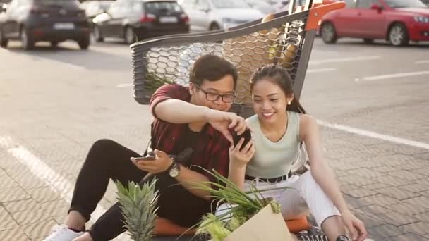 Encantador joven pareja asiática sentada en el suelo cerca de su carrito de mercado y el uso de teléfonos inteligentes — Vídeo de stock