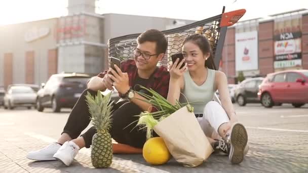 Boldog jó kinézetű ázsiai férfi és nő ül az aszfalt közelében piac trolibusz és használja a telefonok nézni vicces képek — Stock videók