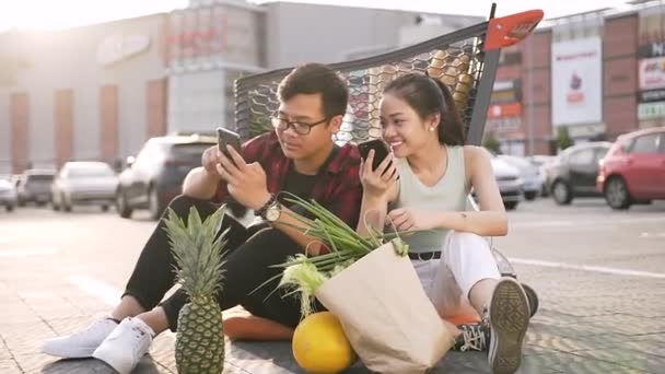 Schöne junge asiatische Mann und Frau in lässiger Kleidung sitzen auf dem Boden vor dem Supermarkt und beobachten auf ihren Handys lustige Fotos — Stockvideo