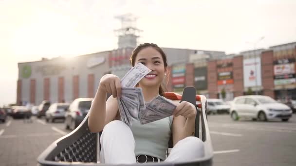 Vonzó képet mosolyogva elégedett ázsiai nő szétszórt pénz, ül a bevásárlókocsin — Stock videók