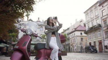 Şık güneş gözlüğü büyüleyici gülümseyen kadın kamera fotoğraf çekmek ve modern scooter üzerinde oturan