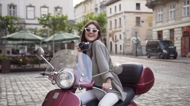 Bella giovane donna in occhiali alla moda con fotocamera vintage e seduto sul moderno scooter in mezzo alla strada della città — Video Stock