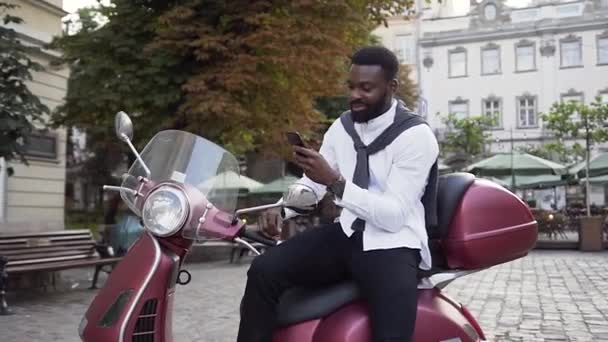 Attraktive bärtige afrikanisch-amerikanische Mann sitzt auf dem neuen Roller und benutzt sein Handy auf dem Hintergrund der schönen städtischen Straße — Stockvideo