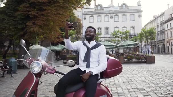 Привлекательный веселый африканский американец, сидящий на модном скутере и делающий селфи рядом с замечательными зданиями — стоковое видео