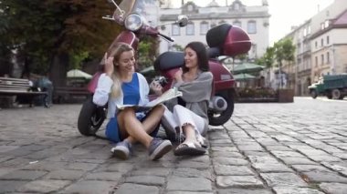 Güzel scooter yakın kaldırımda oturan ve şehir haritası bakarak Pretty mutlu genç kadınlar