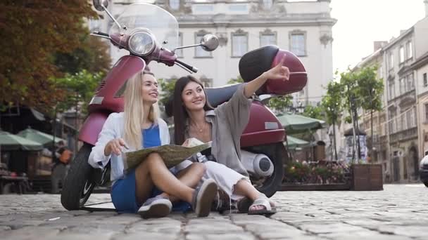 Вероятные веселые подруги сидят на асфальте рядом со скутером и смотрят на карту города, чтобы найти достопримечательности — стоковое видео