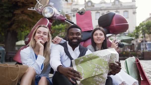 Jovens felizes sentados no chão perto de scooter e usando o mapa da cidade para saber a direção certa no fundo de belos edifícios — Vídeo de Stock