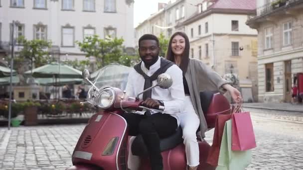 Sympathische fröhliche Mischlingspaare, die auf dem Roller sitzen und Mädchen, die Einkäufe vor dem Hintergrund der Stadtarchitektur in den Händen halten — Stockvideo