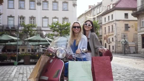Прекрасные веселые подруги в модных солнцезащитных очках, сидящие на скутере с покупками в центре городской площади — стоковое видео
