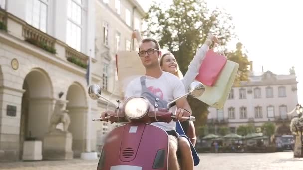 Atractiva pareja joven montando en scooter en la calle urbana y la mujer feliz levantó las manos con las compras — Vídeo de stock